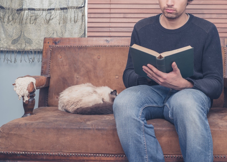 A young man is sitting on a sofa with a cat and is reading a big book