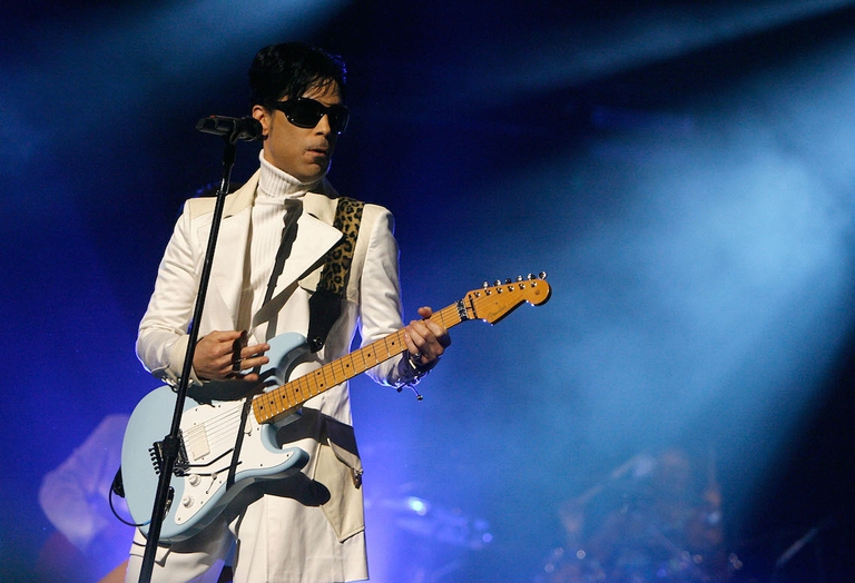 Prince sul palco vestito di bianco con la sua chitarra.